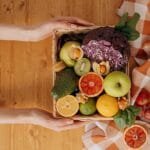 Vibrant basket of fresh fruits and vegetables with hands holding on a wooden table.